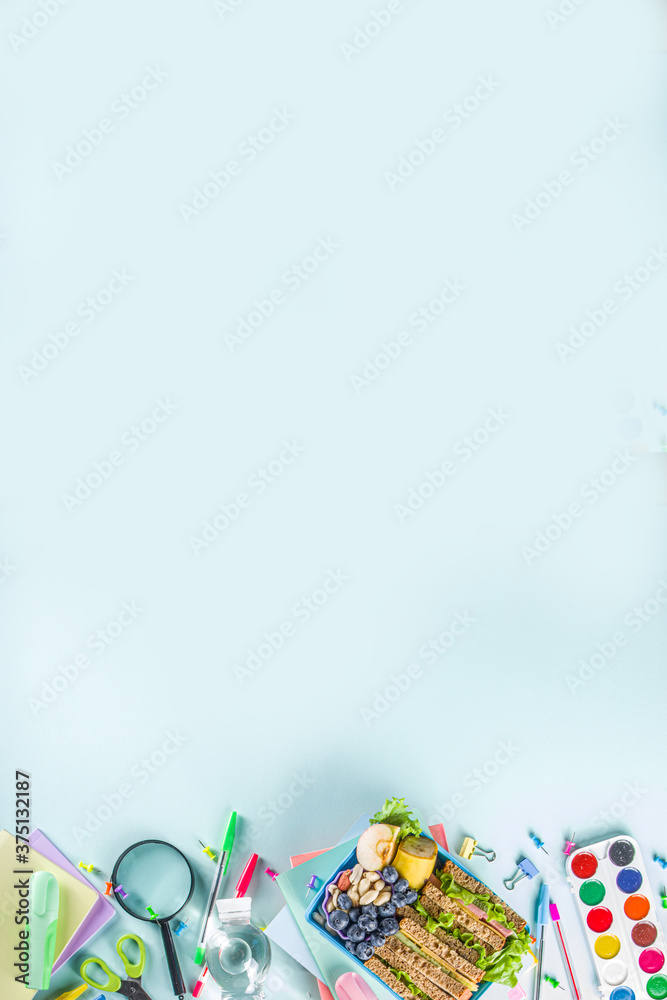 Fototapeta premium Healthy children school lunch box: sandwich, vegetables ,fruit and mineral water bottle, with school supplies on, on light blue background copy space