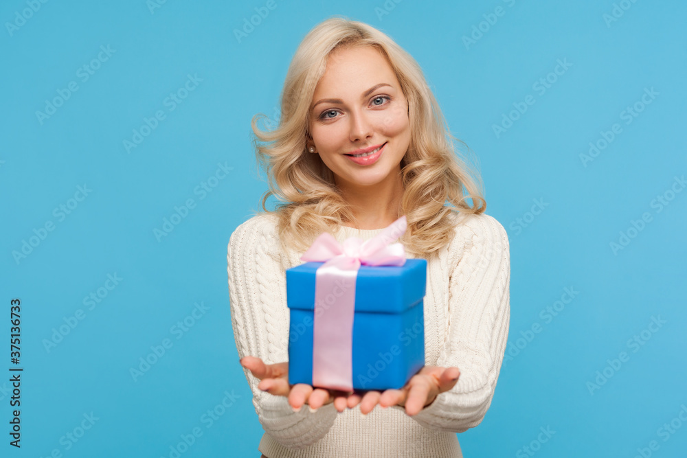 Cute positive blond woman in knitted sweater holding in hands blue giftbox with ribbon bow, making present, surprise. Indoor studio shot isolated on blue background Cute positive blond woman in knitted sweater holding in hands blue giftbox with ribbon bow, making present, surprise. Indoor studio shot isolated on blue background