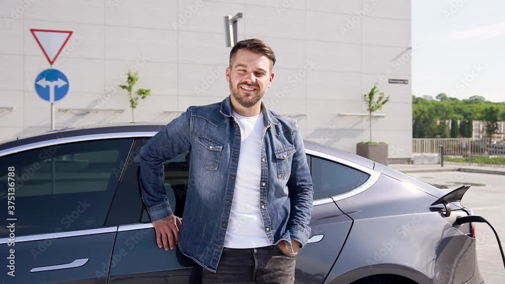 Handsome smiling young bearded man posing on camera near his electric car that charging on specially-equipped charging station Handsome smiling young bearded man posing on camera near his electric car that charging on specially-equipped charging station