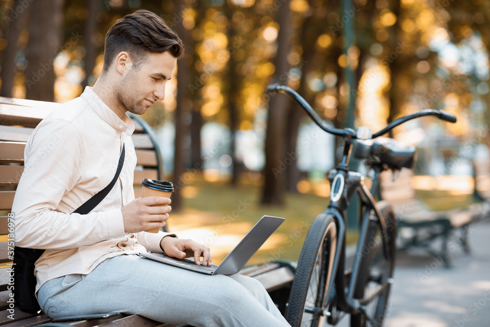 Eco transport and work in city. Young man working on laptop and drinking coffee on bench Eco transport and work in city. Young man working on laptop and drinking coffee on bench