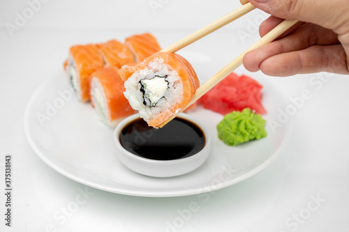 take a philadelphia maki rolls with bamboo chopsticks and put in into soy sauce. pink ginger, wasabi on white plate, asian food, japanese cuisine background