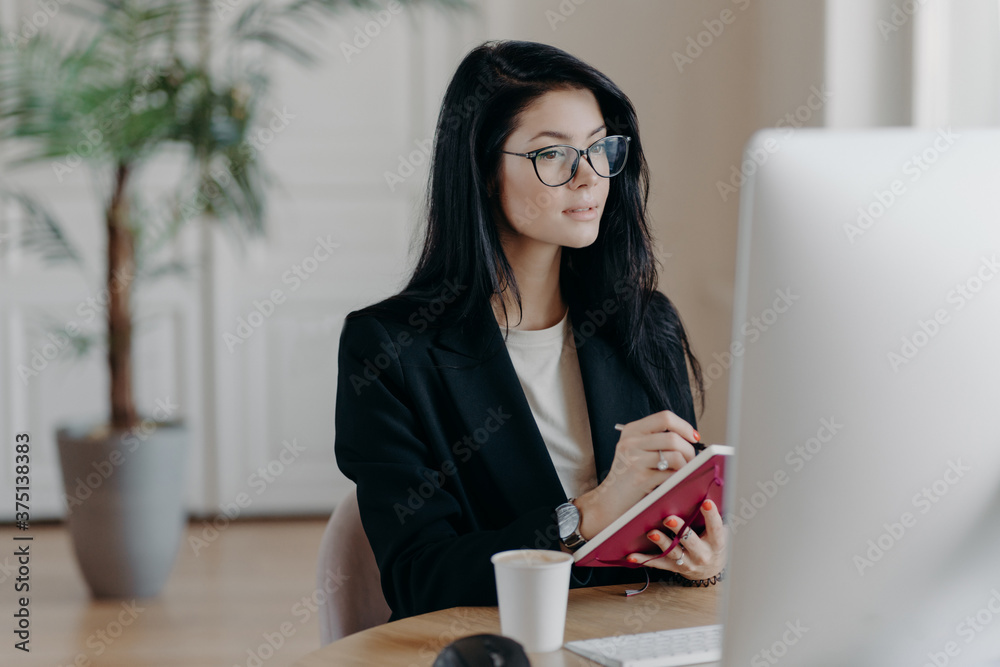 Indoor shot of prosperous businesswoman makes notes in notepad, concentrated in computer screen, watches training video how to start own business, poses at desktop with coffee cup, makes report Indoor shot of prosperous businesswoman makes notes in notepad, concentrated in computer screen, watches training video how to start own business, poses at desktop with coffee cup, makes report