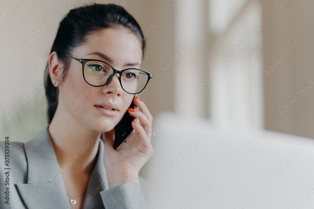 Close up shot of pleasant looking brunette European woman wears transparent glasses, focused in laptop screen, has telephone conversation during remote work, works on project. Technology concept