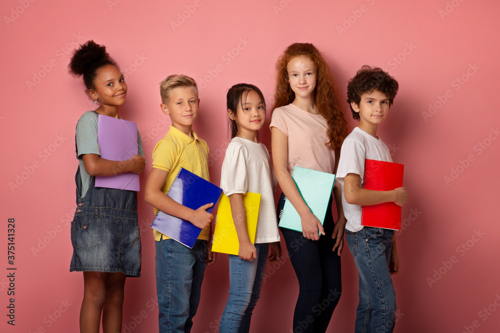 Cheerful multiethnic schoolchildren standing with notebooks in their hands over pink background Cheerful multiethnic schoolchildren standing with notebooks in their hands over pink background