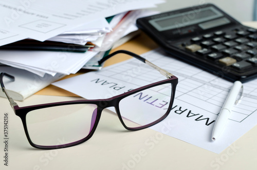 Closeup of glasses, blank paper for marketing plan, pile of documents, calculator on the desk