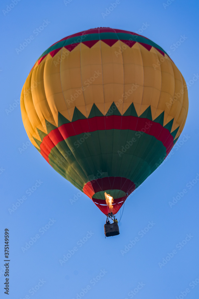 Fototapeta premium A bright multicolored hot air balloon with an expansive burner flame soars up against a background of blue sky. Colorful balloon watercolor painting. Close-up.