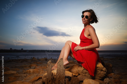 Brunette adult woman in red dress and sunglasses posing in salt lake on beautiful sunset background
