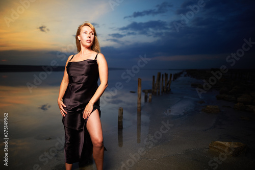 Blonde adult woman in beautiful elegance dress standing in salt lake on sunset background 