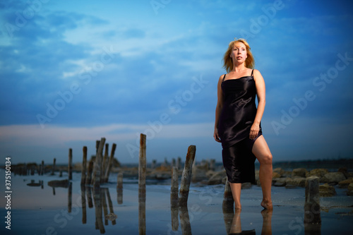 Blonde adult woman in beautiful elegance dress standing in salt lake on sunset background 