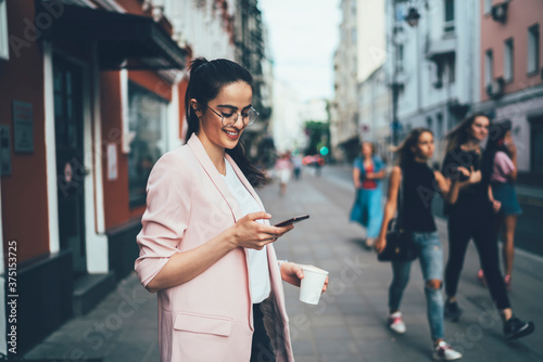 Joyful hipster girl enjoying network browsing during leisure time in urban town, happy Spanish blogger messaging with friends via modern smartphone using 4g wireless internet for web booking