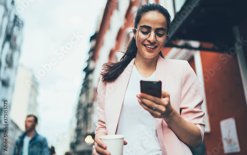 Joyful hipster girl enjoying network browsing during leisure time in urban town, happy Spanish blogger messaging with friends via modern smartphone using 4g wireless internet for web booking