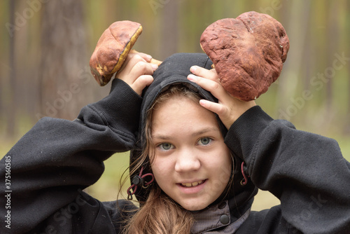Leinwand Poster Cheerful girl in hood holds in her hands large boletus above her head instead of horns in the forest