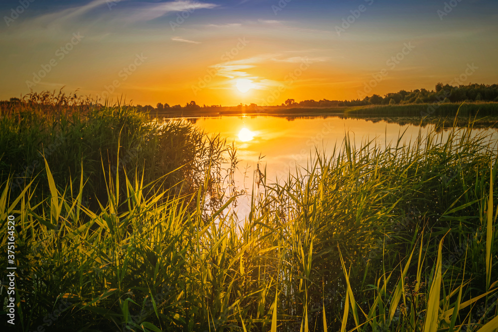 Scenic view at beautiful spring sunset with reflection on a shiny lake with green bushes, grass, golden sun rays, calm water ,deep blue cloudy sky and glow on a background, spring evening landscape