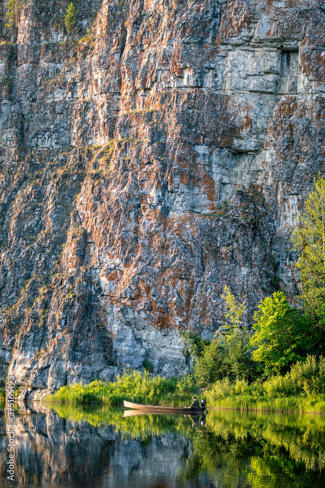 Obraz premium A picturesque landscape with rocks along the river and a fisherman in a wooden boat in the summer season. Ural, Chusovaya river.
