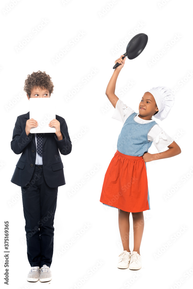 african american kid pretending housewife and holding frying pan near boy in suit covering face with digital tablet isolated on white african american kid pretending housewife and holding frying pan near boy in suit covering face with digital tablet isolated on white