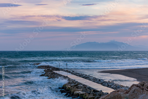Landscape of a breakwater on a beach with waves, mountains in the background and a sunset sky in autumn.