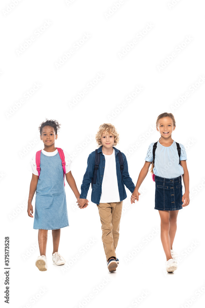 Obraz premium curly schoolboy holding hands with multicultural schoolgirls and walking isolated on white