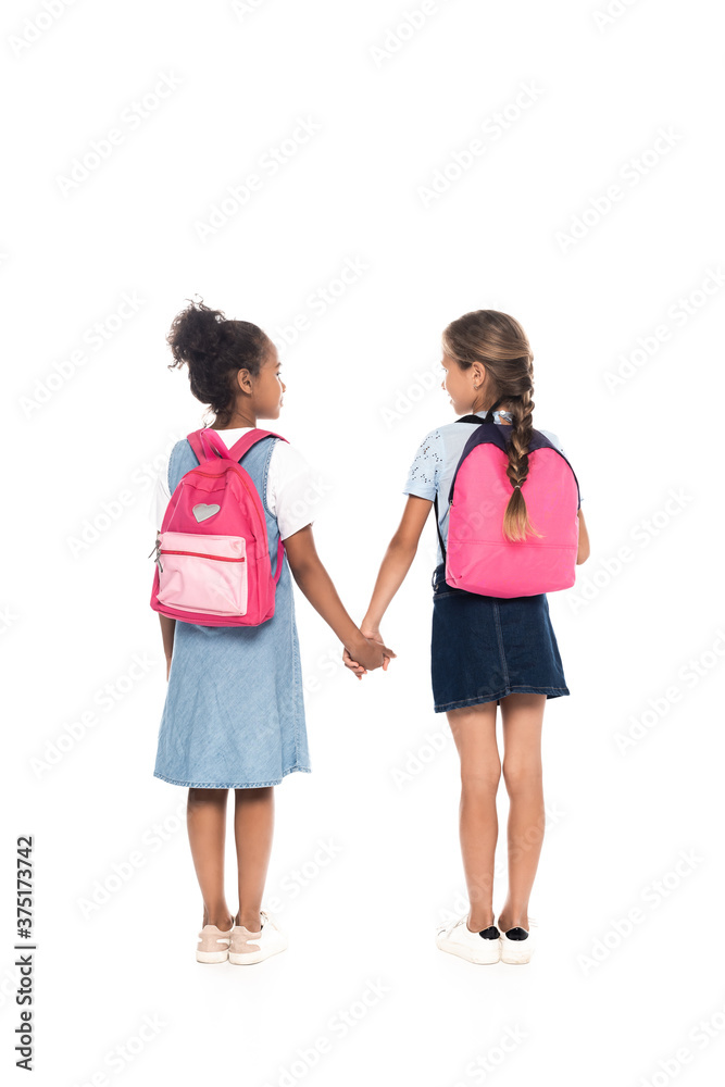 back view of multicultural schoolgirls with backpacks holding hands and standing isolated on white back view of multicultural schoolgirls with backpacks holding hands and standing isolated on white