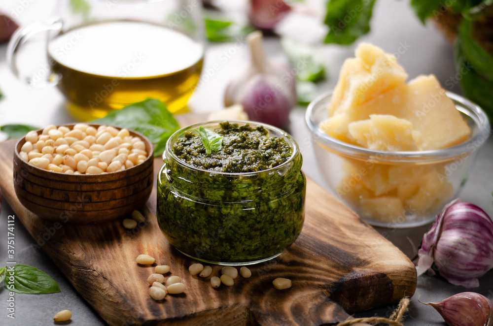 Glass jar with genovese pesto sauce on cutting board with basil leaves, pine nuts, parmesan cheese, olive oil and garlic aside