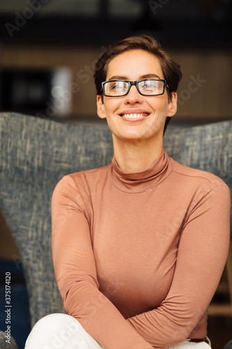 Close up portrait of a smiling woman wearing eyeglasses in an apartment. Positive female in casuals sitting on armchair. photo