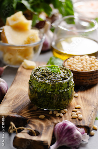 Glass jar with genovese pesto sauce on cutting board with basil leaves, pine nuts, parmesan cheese, olive oil and garlic aside