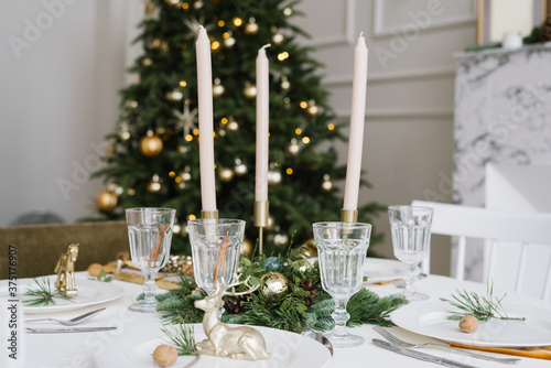 Beautiful glass glasses, candles in the decor of the festive table. Christmas table setting on the background of a Christmas tree