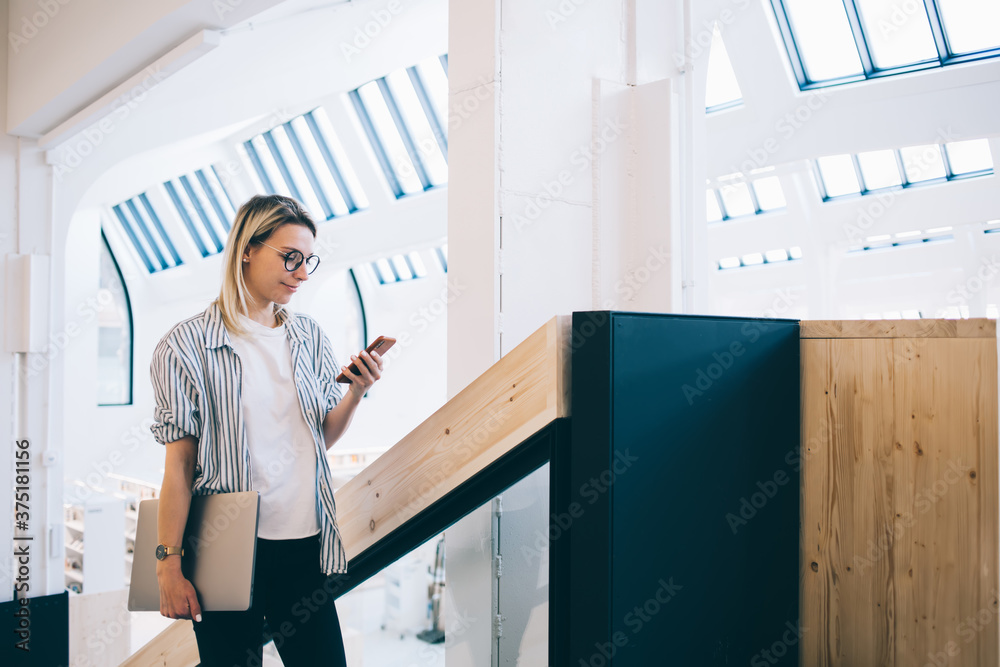 Young freelancer with laptop using smartphone on staircase