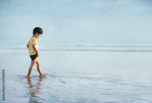 A boy walking on the shallow water at the beach as the waves calm in the morning. The surrounding atmosphere is filled with the same tone of blue in both the sky and the sea.