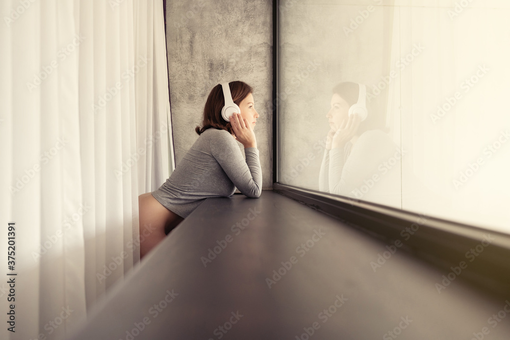 Portrait of beautiful сaucasian young woman in headphones dreaming, enjoying listening music standing and looking at big panoramic window of apartments. Portrait of beautiful сaucasian young woman in headphones dreaming, enjoying listening music standing and looking at big panoramic window of apartments.