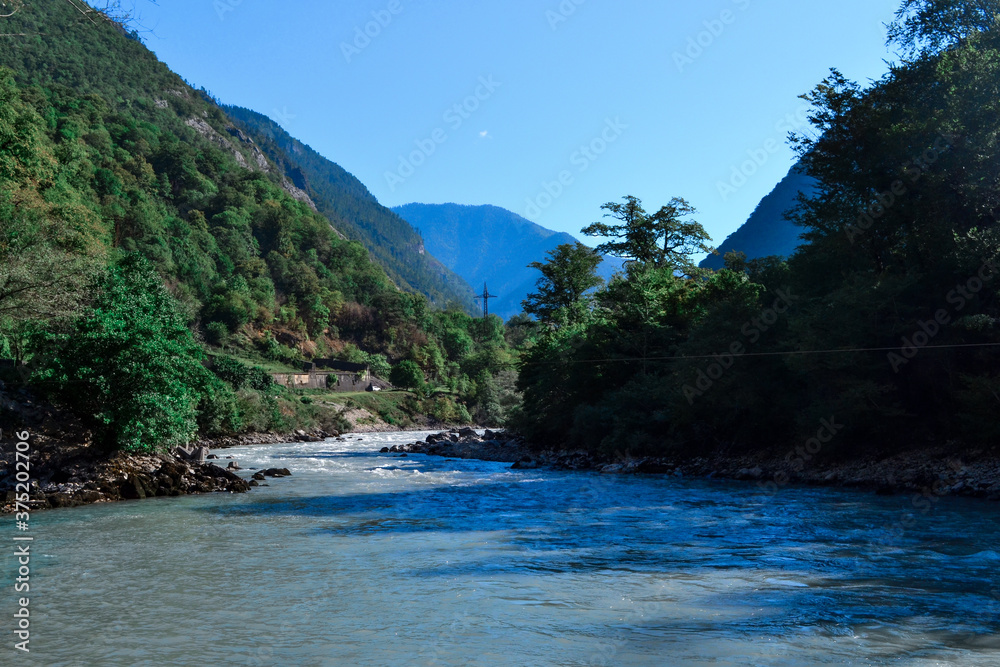 A mountainous blue clear river flows between hills and big mountains covered with green trees and bushes