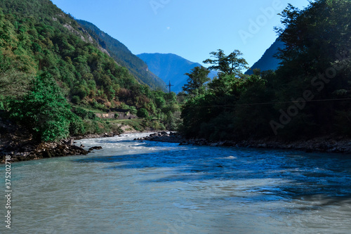A mountainous blue river flows between hills and mountains covered with green trees and bushes © SymbiosisArtmedia