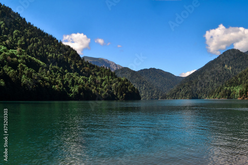 Beautiful blue clear emerald Lake Ritsa in Abkhazia glowing in the sun near the middle of trees green high mountains, blue sky and clouds back