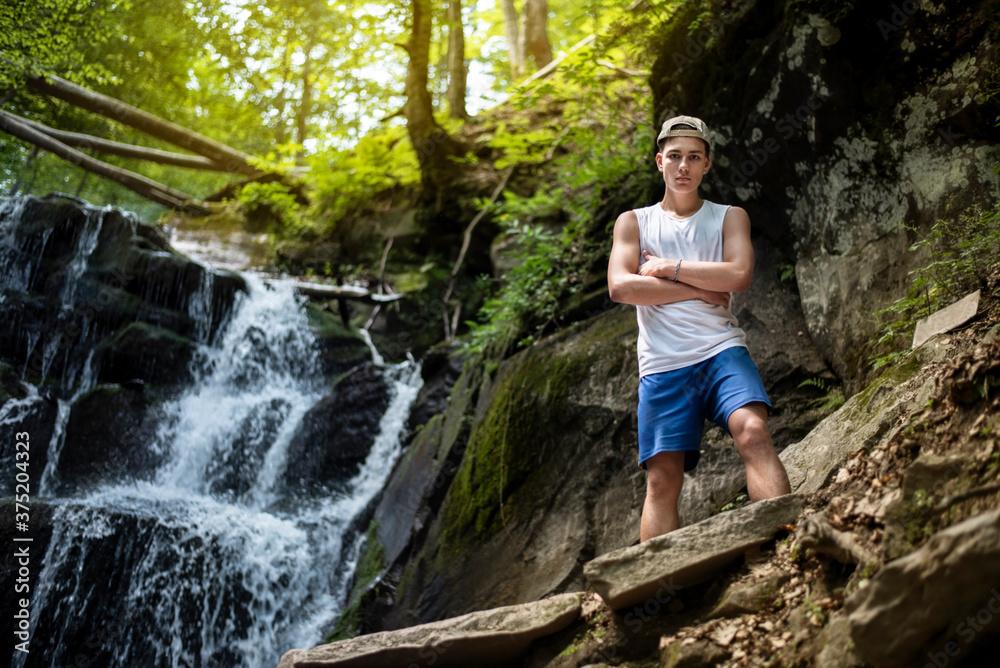 Obraz premium Handsome teenage boy is gaining strength and resting near a waterfall in the mountains, the guy likes to travel through the woods to places of interest