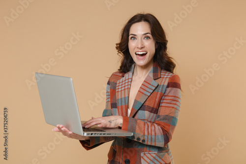 Excited funny young brunette woman 20s wearing casual checkered jacket posing standing holding in hands working on laptop pc computer isolated on pastel beige colour background, studio portrait.