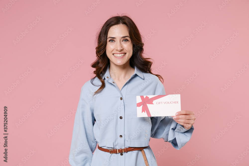 Smiling beautiful young brunette woman 20s wearing casual blue shirt dress posing standing holding in hand gift certificate looking camera isolated on pastel pink colour background, studio portrait. Smiling beautiful young brunette woman 20s wearing casual blue shirt dress posing standing holding in hand gift certificate looking camera isolated on pastel pink colour background, studio portrait.
