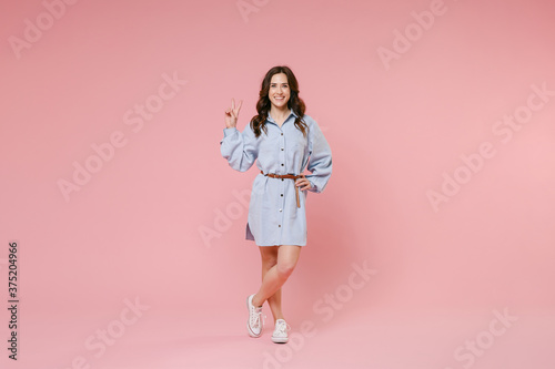 Full length portrait of smiling funny young brunette woman 20s wearing casual blue shirt dress posing standing showing victory sign looking camera isolated on pastel pink colour background in studio. © ViDi Studio