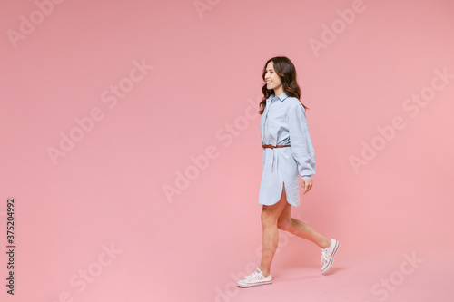 Full length side view portrait of smiling cheerful young brunette woman 20s wearing casual blue shirt dress posing walking going looking aside isolated on pastel pink colour background in studio.