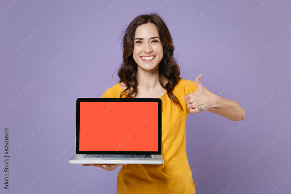 Smiling young brunette woman 20s in yellow t-shirt hold laptop pc computer with blank empty screen mock up copy space showing thumb up isolated on pastel violet colour background, studio portrait. Smiling young brunette woman 20s in yellow t-shirt hold laptop pc computer with blank empty screen mock up copy space showing thumb up isolated on pastel violet colour background, studio portrait.