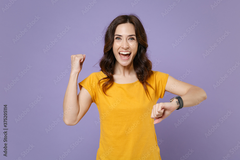 Happy joyful young brunette woman 20s wearing basic yellow t-shirt smart watch on hand posing standing doing winner gesture looking camera isolated on pastel violet colour background, studio portrait. Happy joyful young brunette woman 20s wearing basic yellow t-shirt smart watch on hand posing standing doing winner gesture looking camera isolated on pastel violet colour background, studio portrait.