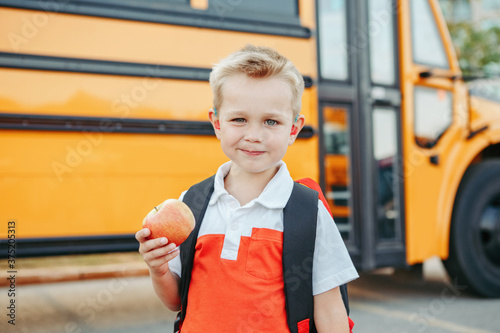 Caucasian boy student with backpack near yellow bus on first September day. Child kid eating apple fruit at school yard outdoors. Education and back to school in Autumn Fall.