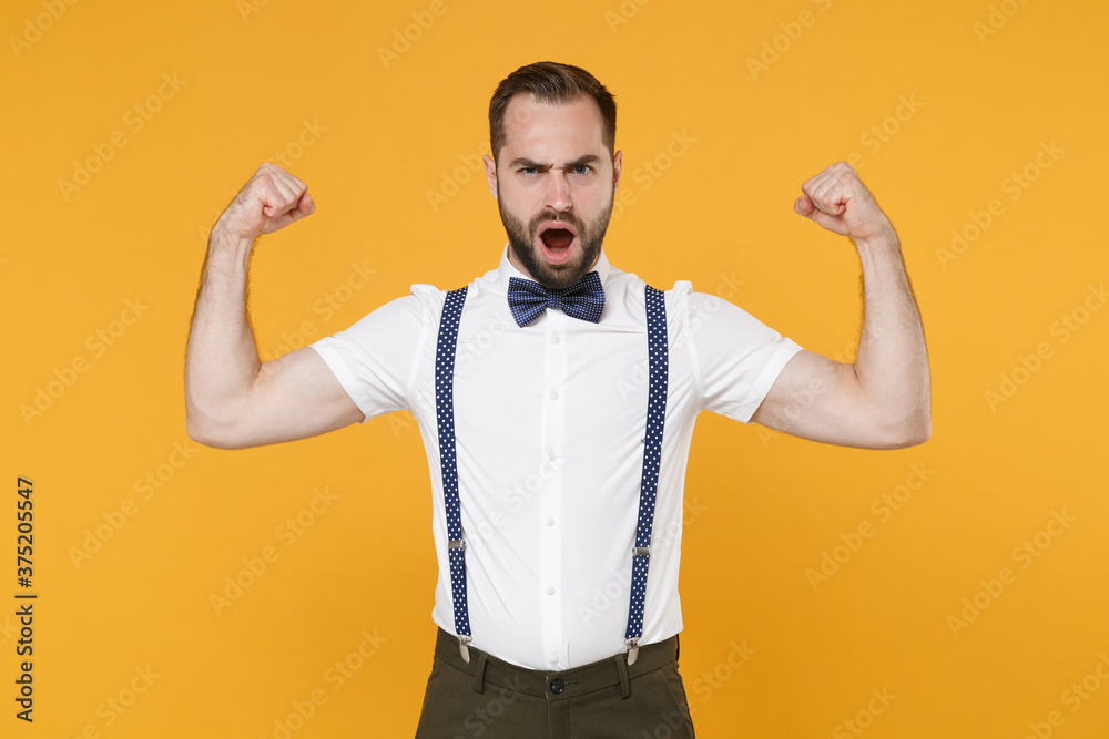Shocked strong young bearded man 20s wearing white shirt bow-tie suspender posing showing biceps muscles keeping mouth open looking camera isolated on bright yellow color background, studio portrait. Shocked strong young bearded man 20s wearing white shirt bow-tie suspender posing showing biceps muscles keeping mouth open looking camera isolated on bright yellow color background, studio portrait.