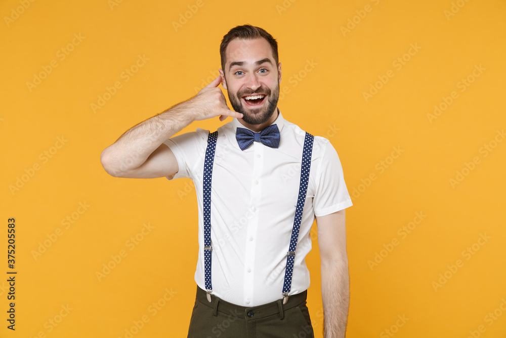 Cheerful funny young bearded man 20s wearing white shirt bow-tie suspender posing doing phone gesture like says call me back looking camera isolated on bright yellow color background studio portrait. Cheerful funny young bearded man 20s wearing white shirt bow-tie suspender posing doing phone gesture like says call me back looking camera isolated on bright yellow color background studio portrait.