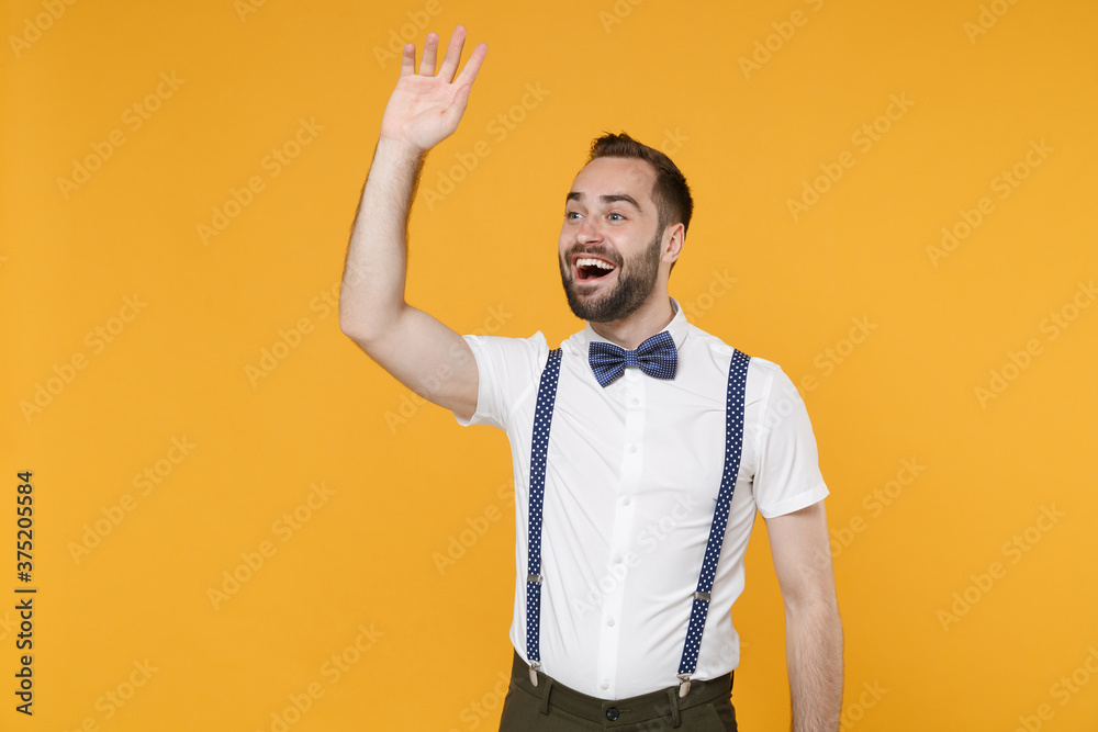 Excited cheerful young bearded man 20s in white shirt bow-tie suspender posing waving and greeting with hand as notices someone looking aside isolated on yellow color wall background studio portrait. Excited cheerful young bearded man 20s in white shirt bow-tie suspender posing waving and greeting with hand as notices someone looking aside isolated on yellow color wall background studio portrait.