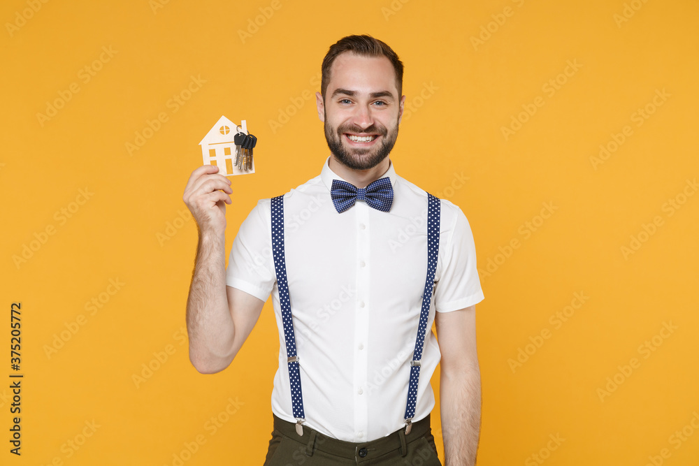 Smiling young bearded man 20s wearing white shirt bow-tie suspender posing standing hold in hand house bunch of keys looking camera isolated on bright yellow color wall background studio portrait. Smiling young bearded man 20s wearing white shirt bow-tie suspender posing standing hold in hand house bunch of keys looking camera isolated on bright yellow color wall background studio portrait.