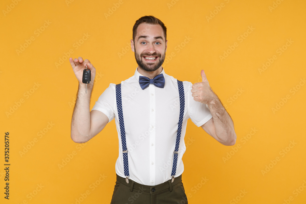 Smiling young bearded man 20s wearing white shirt bow-tie suspender posing standing showing thumb up hold car keys looking camera isolated on bright yellow color wall background studio portrait. Smiling young bearded man 20s wearing white shirt bow-tie suspender posing standing showing thumb up hold car keys looking camera isolated on bright yellow color wall background studio portrait.