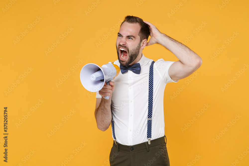 Angy irritated young bearded man 20s wearing white shirt bow-tie suspender posing screaming in megaphone put hand on head looking aside isolated on bright yellow color wall background studio portrait. Angy irritated young bearded man 20s wearing white shirt bow-tie suspender posing screaming in megaphone put hand on head looking aside isolated on bright yellow color wall background studio portrait.