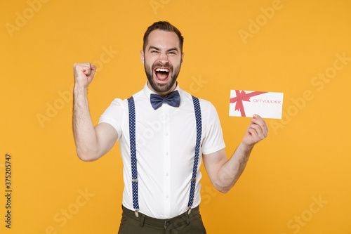 Screaming young bearded man 20s wearing white shirt bow-tie suspender posing standing doing winner gesture hold in hand gift certificate isolated on bright yellow color background studio portrait. © ViDi Studio