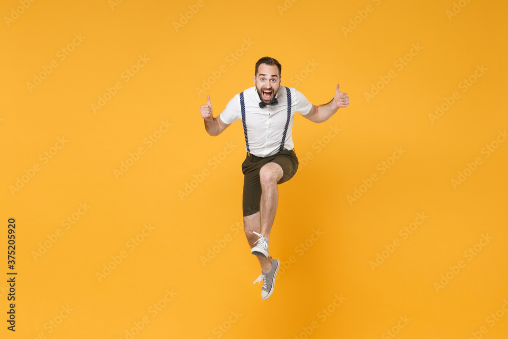 Full length portrait of excited young bearded man 20s wearing white shirt suspender shorts posing jumping showing thumbs up looking camera isolated on bright yellow color wall background studio.