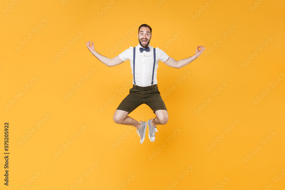 Full length portrait of excited young bearded man 20s wearing white shirt suspender shorts posing jumping hold hands in yoga gesture, relaxing meditating isolated on bright yellow background studio.