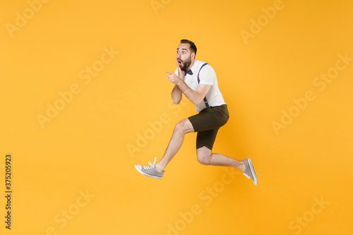 Full length side view portrait of shocked young bearded man 20s in white shirt suspender shorts posing jumping pointing index fingers aside on mock up copy space isolated on yellow background studio.